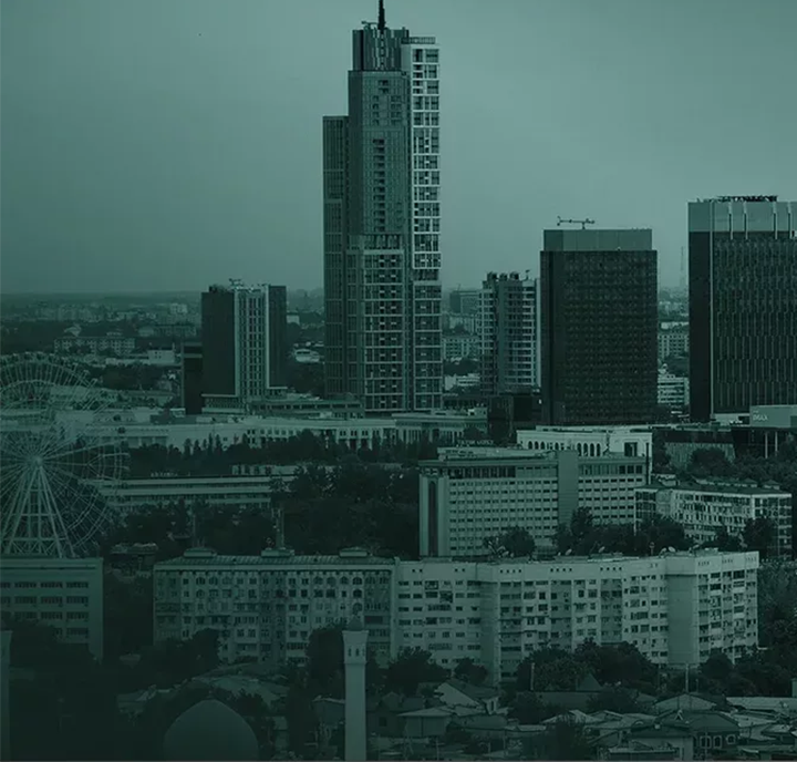 Cityscape view of Almaty, Kazakhstan, with multiple high-rise buildings under a greenish tint. A tall, prominent skyscraper stands centrally, surrounded by shorter buildings. A Ferris wheel is visible to the left, and trees are scattered throughout the urban area. Cloudy sky above.